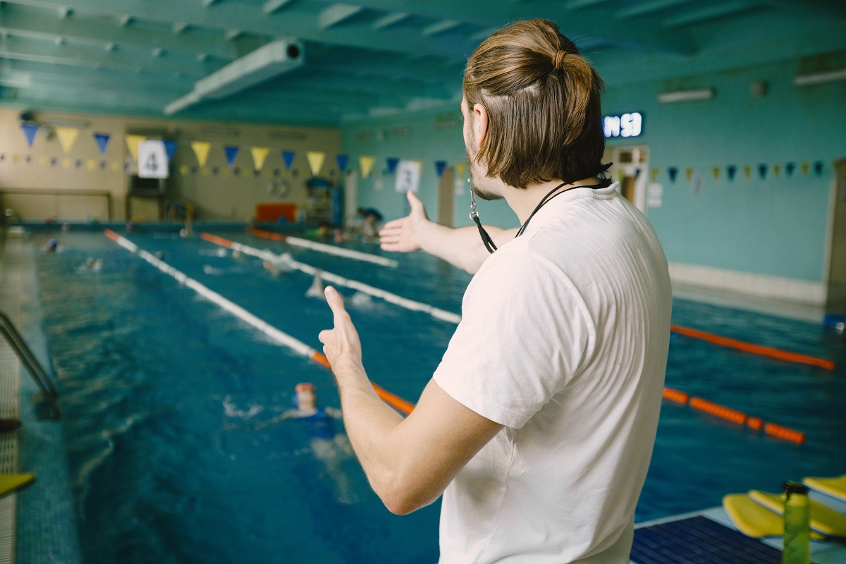 entrenador-natacion-masculino-pie-junto-piscina-mostrando-mano-derecha-corregir-al-estudiante-tecnica-natacion-profesional